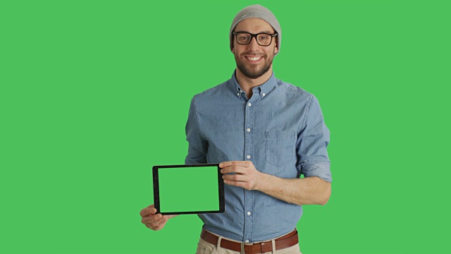 一个微笑的年轻人戴着眼镜展示绿色屏幕的平板电脑的中景。背景中的绿色屏幕。