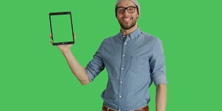 中景一名戴着帽子和眼镜的时尚男子用一只手拿着平板电脑，用另一只手做滑动触摸手势。平板电脑和背景都是绿屏。