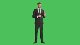 一位英俊的商人用一只手拿着智能手机，用另一只手做滑动触摸手势。平板电脑和背景都是绿屏。视频素材模板下载