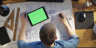 建筑工程师在图纸上的俯视图，与平板电脑和绿屏的比较，也使用桌面电脑。他的桌子上摆满了有用的物品和早晨的阳光。