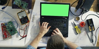 Top View of a Gifted IT技术员类型在他的绿色屏幕笔记本电脑上，而坐在他的办公桌，他被各种技术组件，草稿包围。阳光照在他的桌子上。