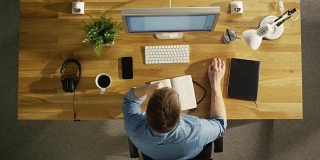 俯视图的一个创造性的设计师的整个工作日的延时。在他的台式电脑上工作，喝咖啡，去吃午饭，在笔记本上写东西和回家。
