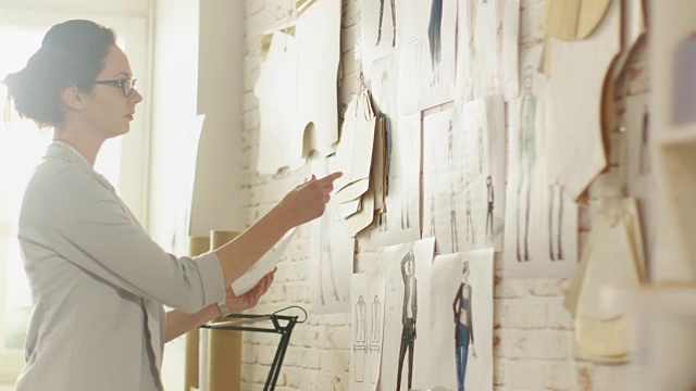 女时装设计师为她的项目选择草图并把它们钉到墙上。她的工作室是Sunny，缝纫机放在桌子上。