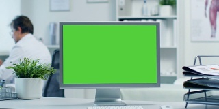 一个绿色屏幕显示器的特写。医生使用文件夹工作，助手通过电话与病人交谈。拍摄于一个明亮和现代的医生的办公室。