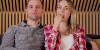 年轻夫妇坐在客厅的沙发上看电视，一边吃薯条