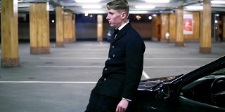 年轻的商人穿着黑色西装，打着领结，坐在停车场的黑色汽车引擎盖前抽烟。