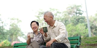 慢镜头快乐的亚洲老夫妇坐在早上的公园喝茶，喝咖啡。交谈并表达爱意