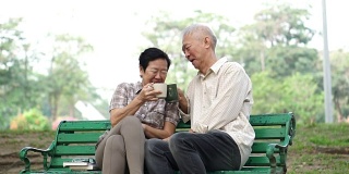 慢镜头快乐的亚洲老夫妇坐在早上的公园喝茶，喝咖啡。交谈并表达爱意