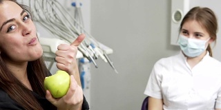 年轻女子咬一个绿苹果在牙科诊所，而坐在附近的牙医后，程序。竖起大拇指。健康的牙齿和健康的微笑。