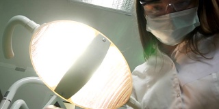 年轻女牙医戴着口罩，带着工具走近。牙科灯照进病人嘴里，站在病人身上，看着相机，牙医的脸