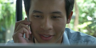 4K:年轻的亚洲商人一边用笔记本电脑一边用手机通话