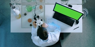 一个科学家在个人电脑上工作的俯视图，绿色的屏幕，观察显微镜和用图表把结果写在文件上。