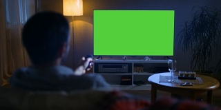 一位中年男子坐在沙发上看大屏电视，他用遥控器切换频道。这是晚上。