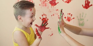 年轻的母亲和她的小男孩在他们的手混合颜色，你留下美丽的手印在墙上。母亲和孩子的概念。Slowmotion。