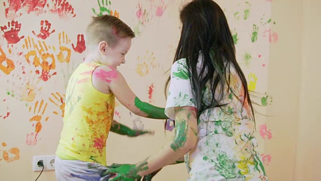 年轻的母亲和她的小男孩在他们的手混合颜色，你留下美丽的手印在墙上。他们的衣服颜色很脏。母亲和孩子的概念。Slowmotion。