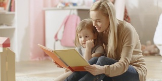 美丽的年轻妈妈读儿童读物给她可爱的小女儿听。儿童的房间是粉红色的和充满了玩具。