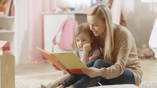 美丽的年轻妈妈读儿童读物给她可爱的小女儿听。儿童的房间是粉红色的和充满了玩具。视频素材模板下载