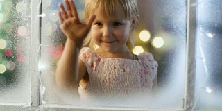 在平安夜可爱的小女孩透过白雪，冰冻的窗户看着和微笑。