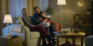 父亲，母亲和小女孩看电视的可能性不大。父亲向他的小女孩解释。他们坐在舒适的客厅里的沙发上。这是晚上。