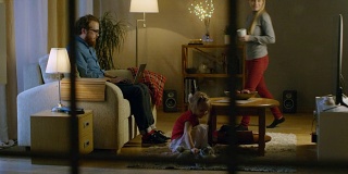 客厅在晚上。父亲坐在沙发上，在用笔记本电脑工作，他的妻子拿着杯子加入他，他们的女儿在他们旁边的地毯上玩软玩具。电视上。