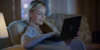 深夜，美丽的女人躺在客厅的沙发上，微笑着在笔记本电脑上打字。她的公寓有大城市的窗户。