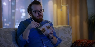 《晚间秀》中，男人坐在客厅的沙发上。他在吃食物盒里的亚洲食物和看电视节目。