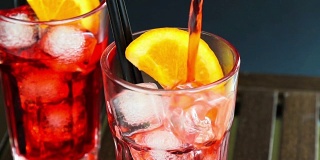 酒保将带有两片橙子和冰块的红色开胃鸡尾酒倒在木桌上