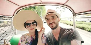 年轻英俊的男子戴着帽子和他的女朋友乘坐泰国传统巴士songteo在智能手机上自拍。一对夫妇在泰国度蜜月
