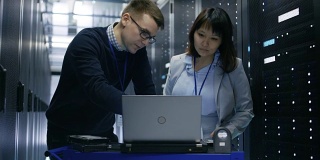 一名男性IT专家向一名女性服务器技术员展示笔记本电脑上的信息。他们正站在数据中心。