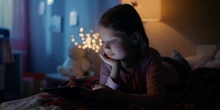 可爱的小女孩晚上在她的房间里，躺在床上使用智能手机。她的夜灯打开了。