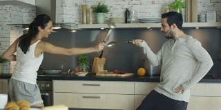 在厨房年轻英俊的夫妇正在与厨房用具击剑。炉子上有东西在锅里煮着。
