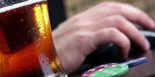 男子喝新鲜啤酒与泡沫在鼠标和电脑键盘附近的赌博芯片背景