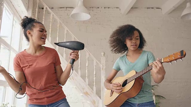 混合种族的年轻有趣的女孩跳舞，与吹风机和弹吉他在床上唱歌。姐妹们有乐趣休闲在卧室在家里