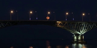满月从桥上升起。延时。俄罗斯萨拉托夫和恩格斯之间的公路桥。伏尔加河。夜晚的车灯和街灯。水中的倒影。一艘货船在桥下航行