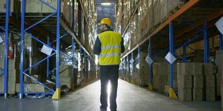 下面是一名仓库工人戴着安全帽走过一排排货架的照片。