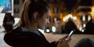 迷人的黑发女子晚上坐在市中心，用触屏技术看智能手机上的照片