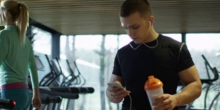 一个年轻的运动员一边用智能手机听音乐，一边在健身房里用跑步机散步。