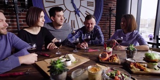 五个朋友拿着红酒坐在餐桌前，在餐厅里吃着不同的零食，开派对。男男女女都在靠窗的咖啡馆里消磨闲暇时光