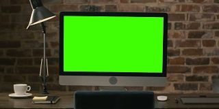 在一间阁楼里，一个绿色屏幕的电脑显示器放在桌子上，旁边是平板电脑、台灯、咖啡杯、笔记本和鼠标。