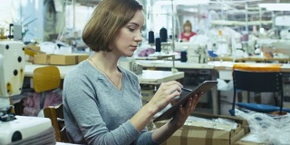 一名年轻女性正坐在服装厂的一张桌子旁，用手写笔在写字板上画素描。