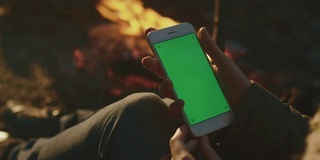 晚上，女孩在篝火旁拿着一个绿色屏幕模型的肖像模式的智能手机。