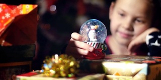 黄昏时分，在色彩鲜艳的纸包装的礼物中，一个金发碧眼的小女孩，头发上系着一个粉红色的蝴蝶结，穿着一件漂亮的裙子，仔细地看着，欣赏着雪花球。里面有一个雪人和一个闪闪发光的圣诞玩具球