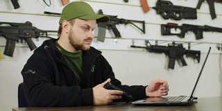 一个枪支贩子在工作。坐在商店的电脑前，在架子的背景上有不同的武器
