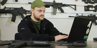 一个枪支商店经理用笔记本电脑工作。展台的背景上有商品的图片