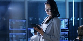 年轻女性公务员在系统控制中心使用平板电脑。在后台，她的同事们在他们的工作空间里有许多显示有价值的数据的显示器。
