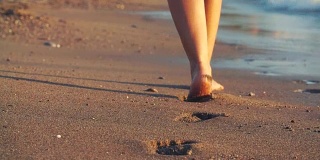 女人美丽的晒黑的腿走在沙滩上。日落时分，一个女孩在海上散步