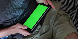 一男子坐在沙发上，手里拿着一台绿色屏幕的平板电脑