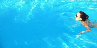 在阳光明媚的夏日里，年轻的黑发女子穿着比基尼和太阳镜在游泳池里游泳