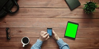 俯视图男性的手在智能手机和数字平板电脑上处理大数据图表，绿色屏幕放在白色的桌子上