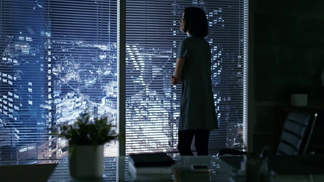 美丽的女商人从她的私人办公室的窗户看向大城市，然后坐在她的办公桌。她的工作空间用暗色调看起来很现代。